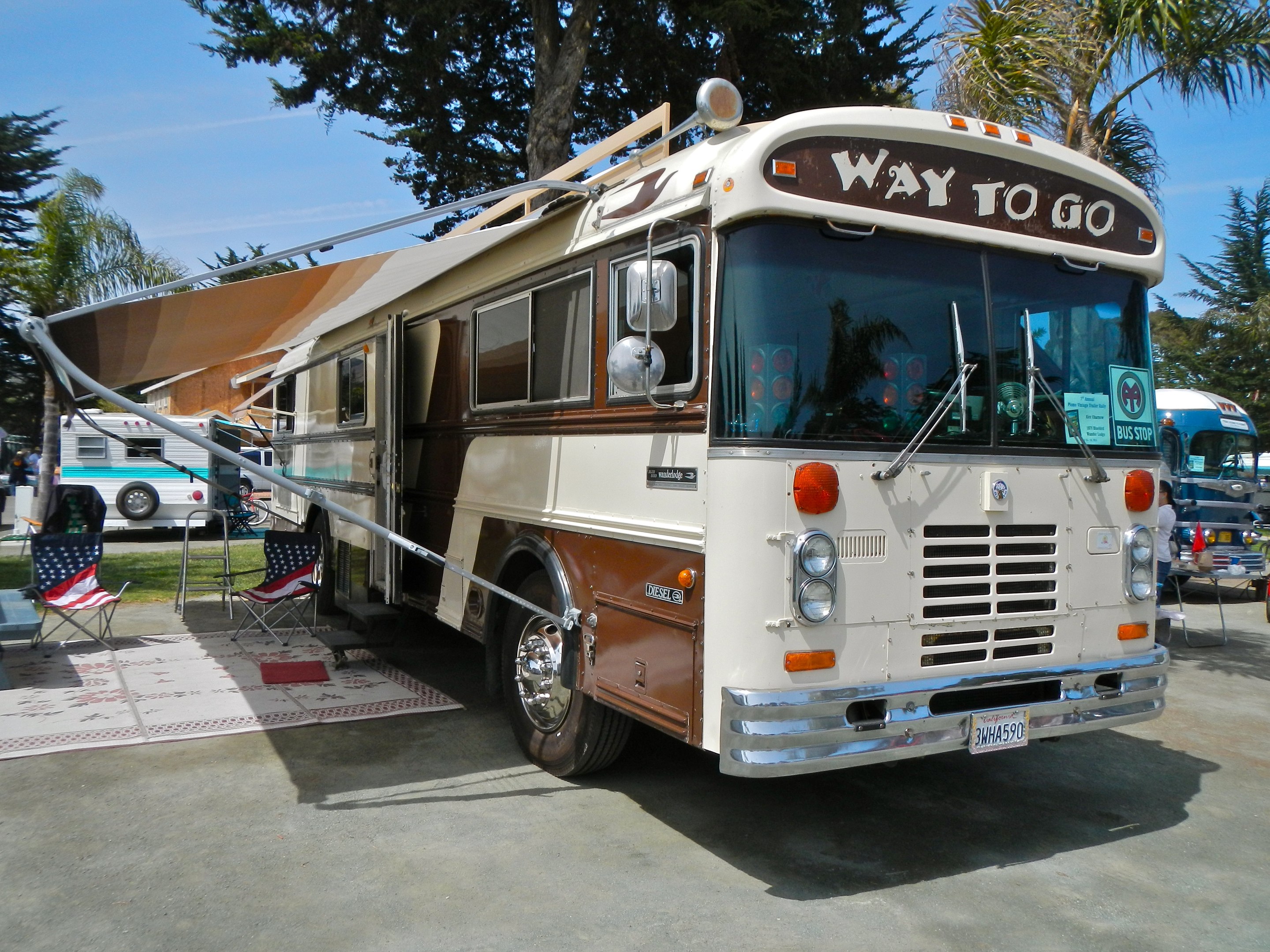 A 1979 Blue Bird Wanderlodge motorhome with Way To Go written on the front parked at an RV show with awning extended and American flag chair outside