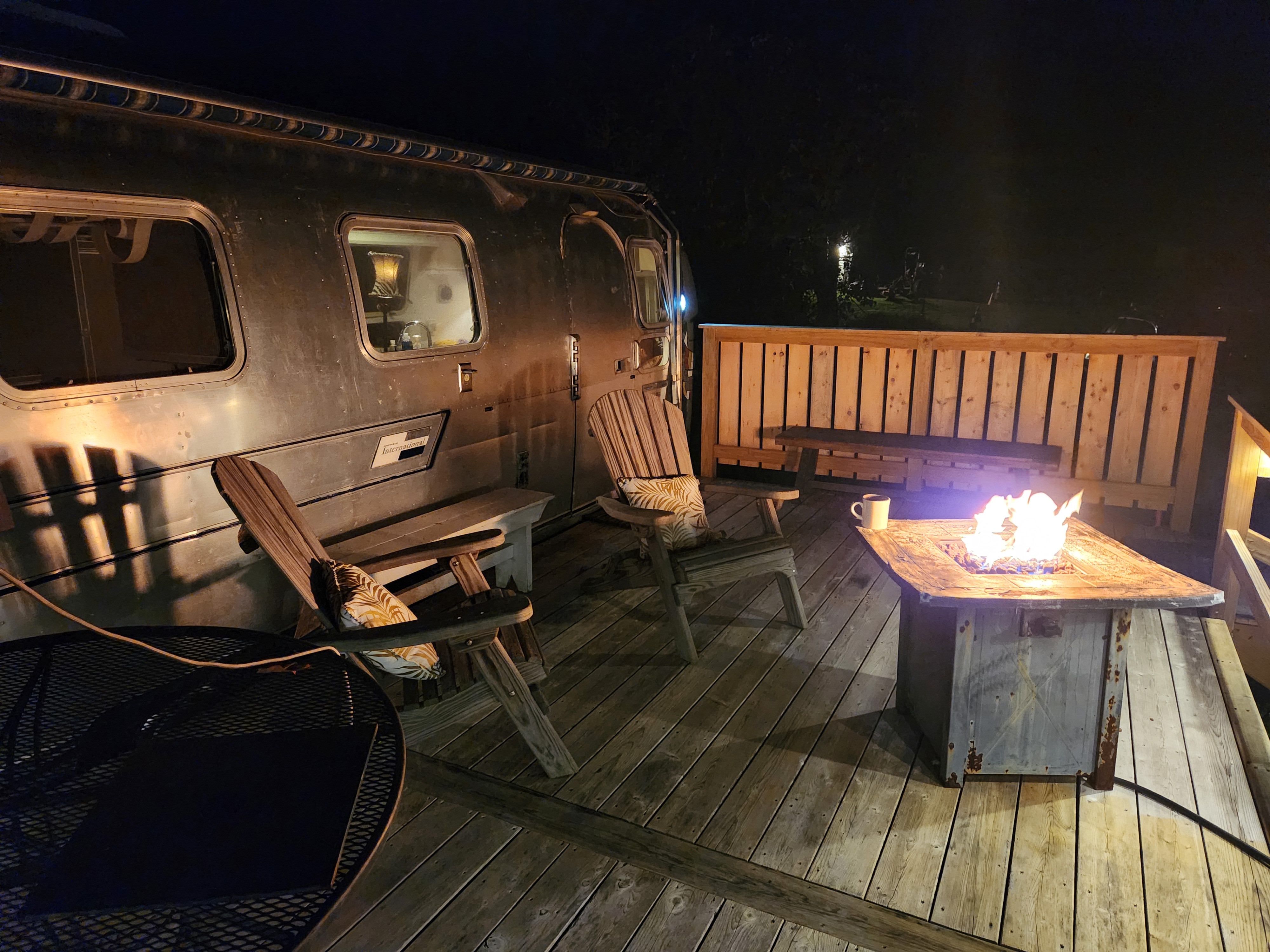 Airstream International travel trailer parked beside a wooden deck with Adirondack chairs and a glowing fire pit table at night