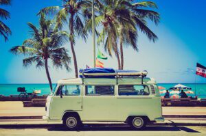 Vintage light blue Volkswagen Type 2 camper van parked at a tropical beach with palm trees and turquoise water representing 1960s and 70s road trip culture