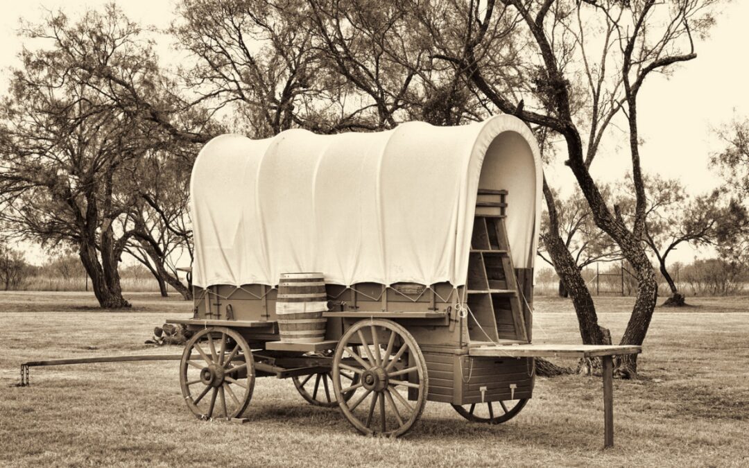 Sepia photo of an old covered chuck wagon with a wooden barrel, parked in a grassy field under bare mesquite trees