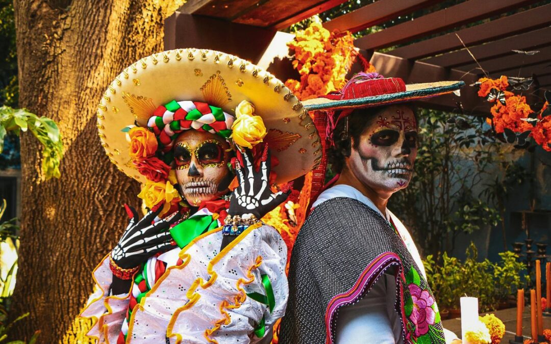 Two people in skull face paint and traditional costumes stand back to back among orange marigolds